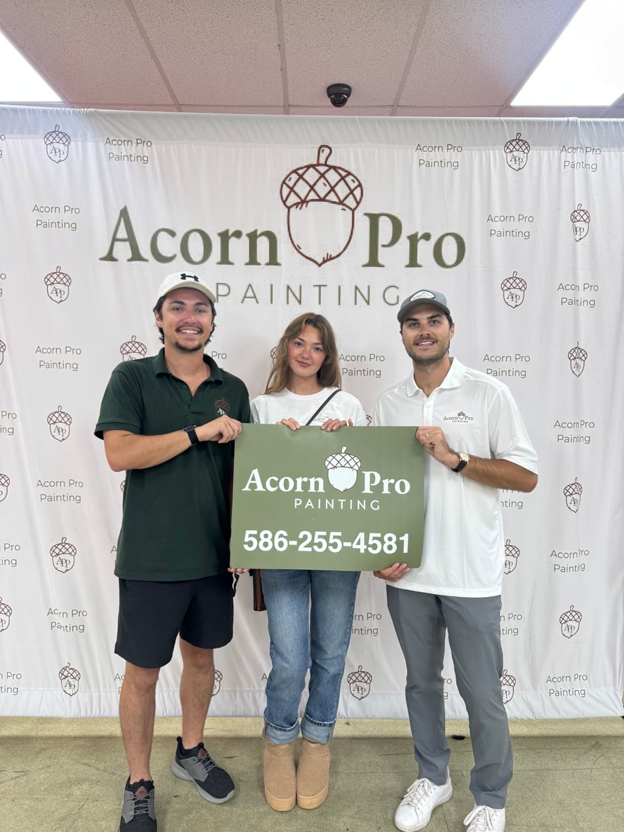 Three Acorn Pro Painting team members holding an Acorn Pro yard sign in front of the branded backdrop